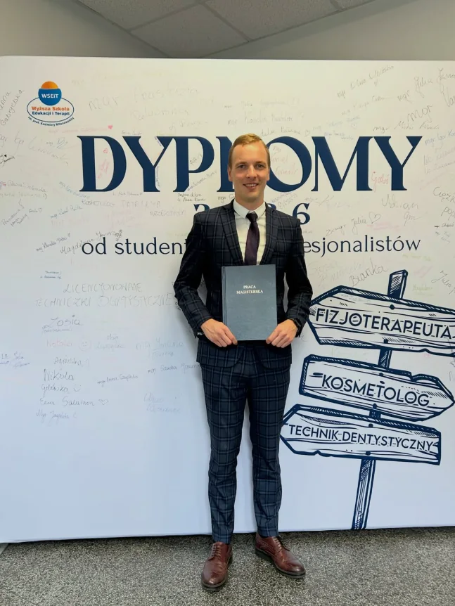 Eryk Myszkowski w garniturze z oprawioną pracą magisterską na ściance „Dyplomy”